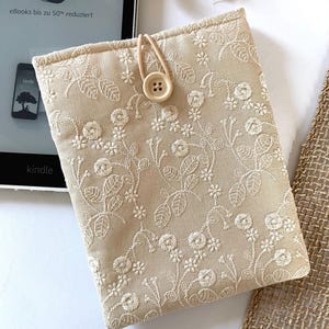 Pu&ograve; includere: Una custodia per e-reader Kindle in lino beige con ricami floreali bianchi e chiusura con bottone marrone.