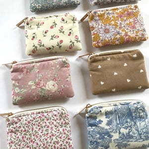 May include: A collection of small, rectangular fabric pouches with zippered closures. The pouches feature various floral and patterned designs in shades of blue, pink, cream, and brown. Each pouch has a gold-toned zipper and a small leather pull.