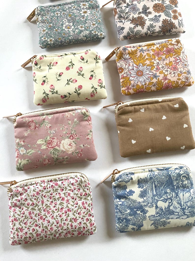 May include: A collection of small, rectangular fabric pouches with zippered closures. The pouches feature various floral and patterned designs in shades of blue, pink, cream, and brown. Each pouch has a gold-toned zipper and a small leather pull.