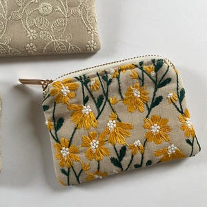 K&ouml;nnte beinhalten: Ein kleiner, beige bestickter M&uuml;nzbeutel mit goldenem Rei&szlig;verschluss und Zipper. Die Geldb&ouml;rse ist mit gelben Blumen und gr&uuml;nen Bl&auml;ttern verziert. Weitere Geldb&ouml;rsen mit floralen Stickereien sind im Hintergrund zu sehen.