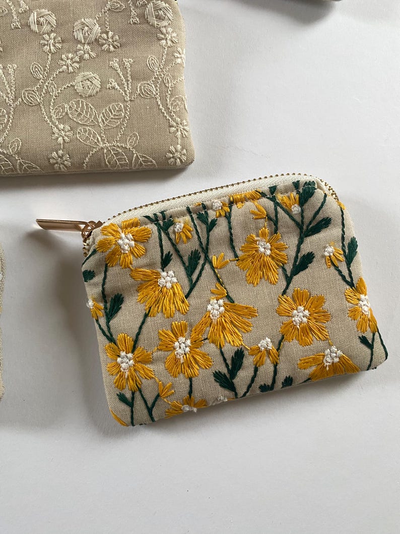 K&ouml;nnte beinhalten: Ein kleiner, beige bestickter M&uuml;nzbeutel mit goldenem Rei&szlig;verschluss und Zipper. Die Geldb&ouml;rse ist mit gelben Blumen und gr&uuml;nen Bl&auml;ttern verziert. Weitere Geldb&ouml;rsen mit floralen Stickereien sind im Hintergrund zu sehen.