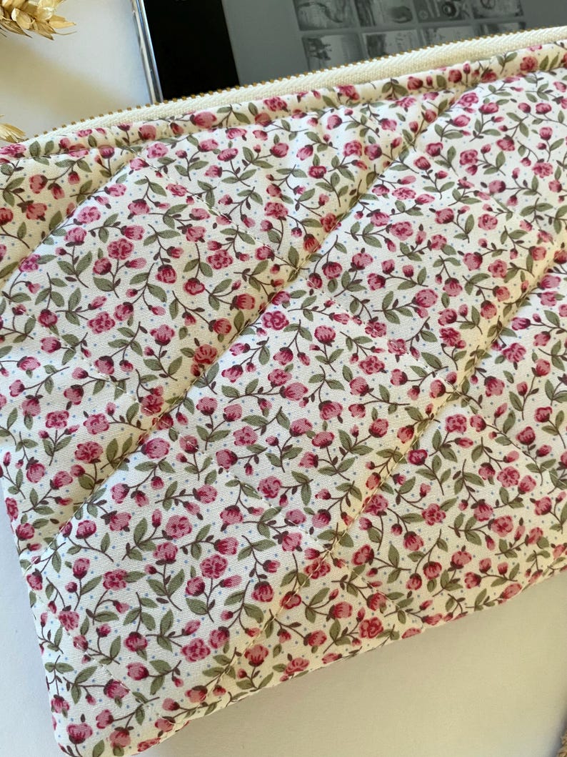 May include: A quilted zippered pouch with a white background and a small pink floral pattern. The pouch is made of a soft fabric and has a zipper closure.