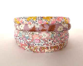 May include: A stack of five fabric-covered headbands featuring various floral patterns in shades of pink, yellow, blue, and white. Each headband has a rounded profile and is wrapped in cotton fabric with distinct botanical prints and colors.