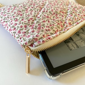 May include: A white zippered pouch with a floral print and a gold zipper. The pouch is open and a black e-reader is inside.