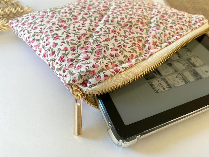 May include: A white zippered pouch with a floral print and a gold zipper. The pouch is open and a black e-reader is inside.