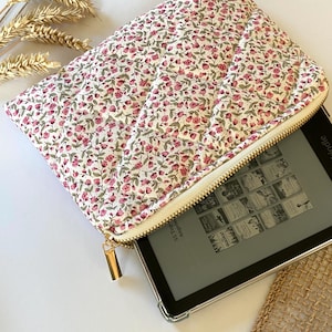 May include: A white quilted zippered pouch with a floral print featuring small pink flowers. The pouch is open and a black e-reader is inside.