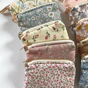 May include: A collection of small, rectangular zippered pouches in various floral patterns. The pouches feature gold-colored zippers and are made of fabric with designs including roses, daisies, and other botanical motifs. The color palette includes pinks, blues, and creams.