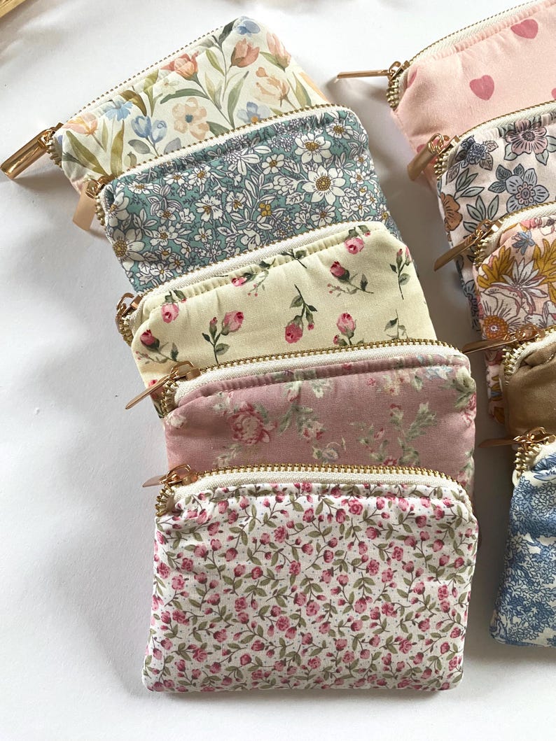 May include: A collection of small, rectangular zippered pouches in various floral patterns. The pouches feature gold-colored zippers and are made of fabric with designs including roses, daisies, and other botanical motifs. The color palette includes pinks, blues, and creams.