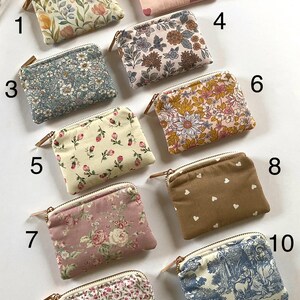 May include: A collection of ten small, rectangular zippered pouches. The pouches feature various floral, heart, and toile patterns in shades of pink, blue, and brown. Each pouch has a gold-toned zipper and a small leather pull.
