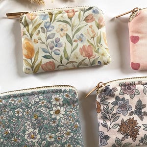 May include: Several small, rectangular coin purses with gold zippers are displayed. The purses feature floral patterns in various colors, including pink, blue, and green. One purse has a pattern of pink hearts. The background is white.