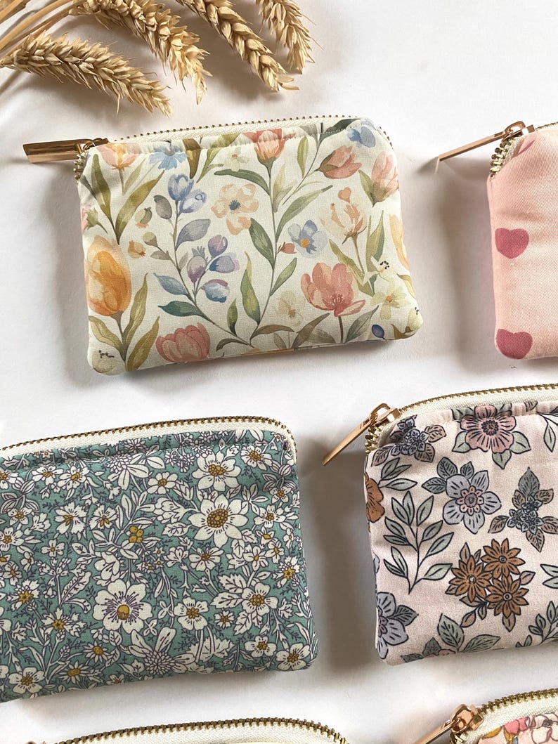 May include: Several small, rectangular coin purses with gold zippers are displayed. The purses feature floral patterns in various colors, including pink, blue, and green. One purse has a pattern of pink hearts. The background is white.