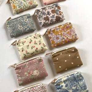 May include: A collection of small, rectangular fabric pouches with zippers. The pouches feature various floral and patterned designs in colors like pink, blue, and brown. Some have heart patterns, while others showcase classic floral prints.