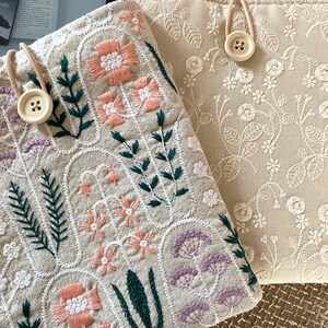 Pu&ograve; includere: Due pochette in lino beige con motivi floreali ricamati. La pochette a sinistra presenta un design pi&ugrave; dettagliato con fiori rosa, verdi e viola. La pochette a destra presenta un design pi&ugrave; semplice con fiori bianchi. Entrambe le pochette hanno una chiusura a bottone.