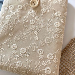 Pu&ograve; includere: Una custodia per tablet in lino beige con motivi floreali ricamati in bianco. La custodia ha un cinturino in pelle marrone e una chiusura a bottone bianco.