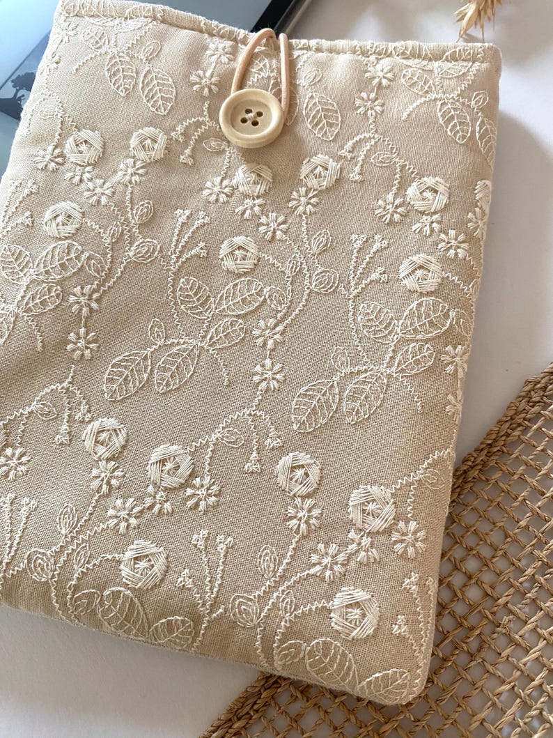 Pu&ograve; includere: Una custodia per tablet in lino beige con motivi floreali ricamati in bianco. La custodia ha un cinturino in pelle marrone e una chiusura a bottone bianco.