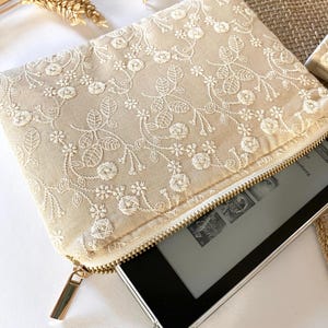 May include: Beige embroidered clutch with a gold zipper, holding an e-reader. The clutch features a floral pattern in white thread. A matching small pouch is visible in the background. The e-reader is black with a silver frame.