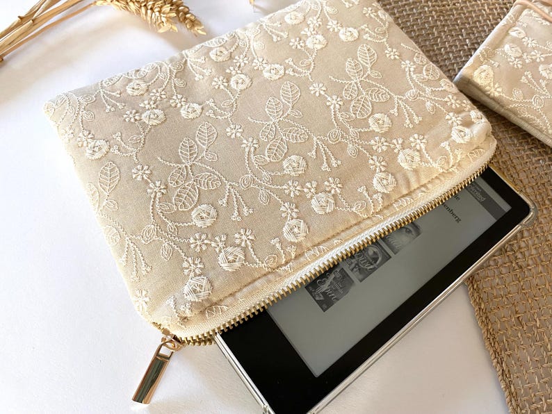 May include: Beige embroidered clutch with a gold zipper, holding an e-reader. The clutch features a floral pattern in white thread. A matching small pouch is visible in the background. The e-reader is black with a silver frame.