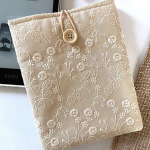 Pu&ograve; includere: Custodia per e-reader Kindle in tessuto beige con ricamo floreale bianco e chiusura con bottone in legno.