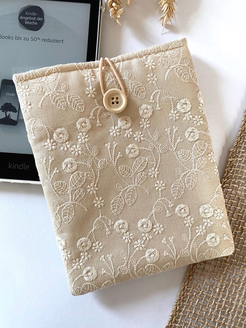 Pu&ograve; includere: Custodia per e-reader Kindle in tessuto beige con ricamo floreale bianco e chiusura con bottone in legno.