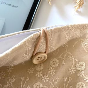 Pu&ograve; includere: Una pochette in tessuto beige con un ricamo floreale bianco. La pochette ha un cinturino elastico marrone e un bottone in legno. Il testo "Kindle Angebot der Woche 50% reduziert" &egrave; visibile sulla pochette.