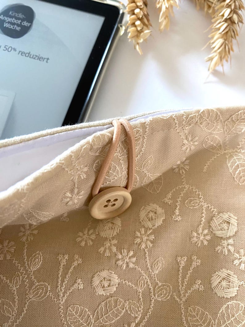 Pu&ograve; includere: Una pochette in tessuto beige con un ricamo floreale bianco. La pochette ha un cinturino elastico marrone e un bottone in legno. Il testo "Kindle Angebot der Woche 50% reduziert" &egrave; visibile sulla pochette.