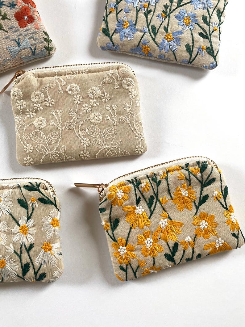 K&ouml;nnte beinhalten: Mehrere kleine, beige bestickte M&uuml;nzb&ouml;rsen mit verschiedenen Blumenmustern. Die Muster zeigen gelbe, blaue, rote und wei&szlig;e Blumen mit gr&uuml;nen Bl&auml;ttern. Jede B&ouml;rse hat einen goldenen Rei&szlig;verschluss und ein beiges Innenfutter.