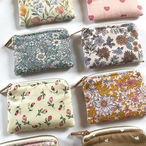 May include: A collection of small, rectangular fabric pouches with zippers. The pouches feature various floral and heart patterns in colors like pink, yellow, blue, and brown. Each pouch has a gold-colored zipper.