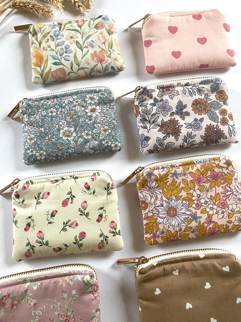 May include: A collection of small, rectangular fabric pouches with zippers. The pouches feature various floral and heart patterns in colors like pink, yellow, blue, and brown. Each pouch has a gold-colored zipper.