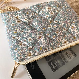 May include: A floral-patterned, quilted pouch in shades of blue, white, and yellow, with a gold zipper. The pouch is open, revealing a black e-reader. The fabric features a repeating pattern of small flowers.