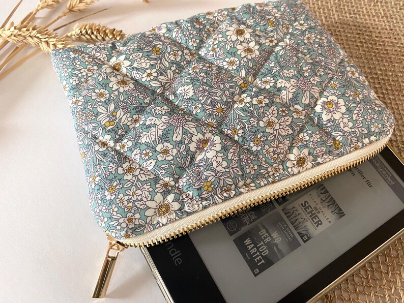 May include: A floral-patterned, quilted pouch in shades of blue, white, and yellow, with a gold zipper. The pouch is open, revealing a black e-reader. The fabric features a repeating pattern of small flowers.