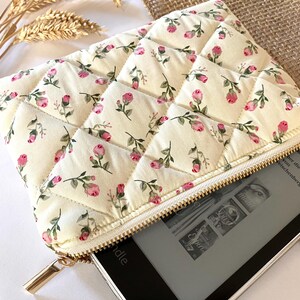 May include: Cream-colored quilted case with a floral pattern of pink roses and green leaves. The case has a gold zipper and is partially open, revealing a black e-reader. The case is designed to protect the e-reader.