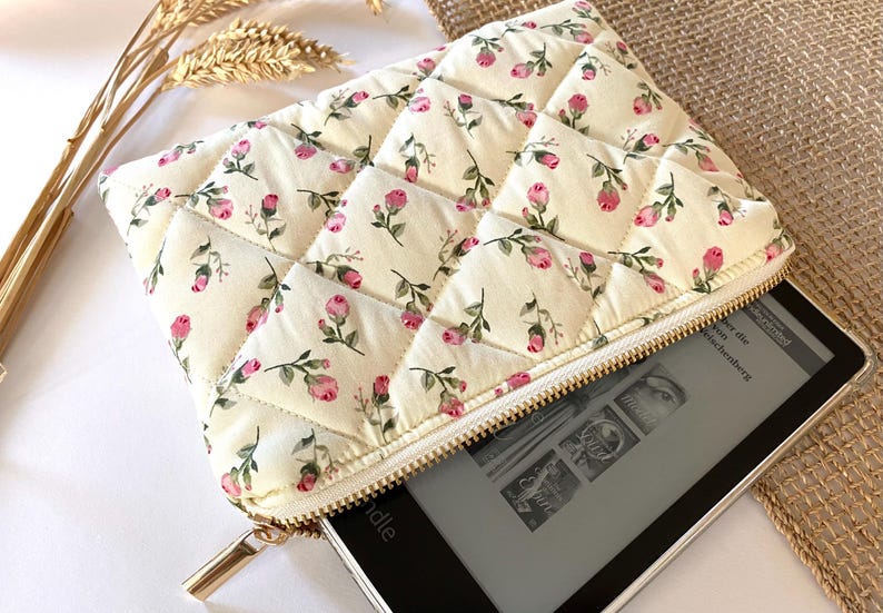 May include: Cream-colored quilted case with a floral pattern of pink roses and green leaves. The case has a gold zipper and is partially open, revealing a black e-reader. The case is designed to protect the e-reader.