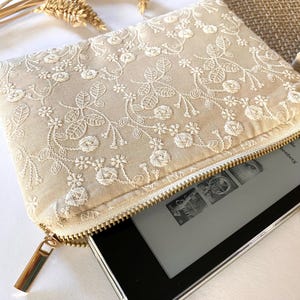 May include: Beige embroidered clutch with a gold zipper, holding an e-reader. The clutch features a floral lace design. The e-reader is partially visible, displaying a book cover. The clutch is rectangular and has a gold zipper pull.