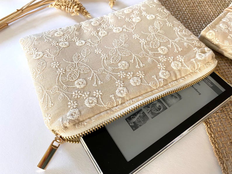 May include: Beige embroidered clutch with a gold zipper, holding an e-reader. The clutch features a floral lace design. The e-reader is partially visible, displaying a book cover. The clutch is rectangular and has a gold zipper pull.