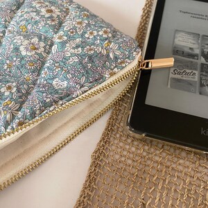 May include: A floral-patterned quilted case with a gold zipper, next to a Kindle e-reader. The case features a light blue background with white, yellow, and purple flowers. The Kindle displays book covers. The case is designed to protect the e-reader.