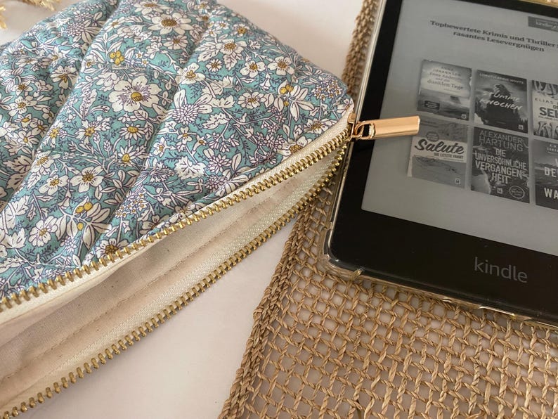 May include: A floral-patterned quilted case with a gold zipper, next to a Kindle e-reader. The case features a light blue background with white, yellow, and purple flowers. The Kindle displays book covers. The case is designed to protect the e-reader.