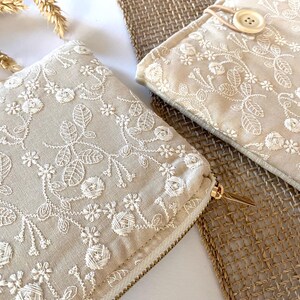 May include: Two beige fabric pouches with intricate white floral embroidery. One pouch has a gold zipper, and the other has a button closure. The pouches are placed on a woven surface, with dried wheat stalks in the background.