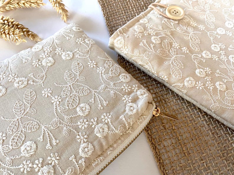 May include: Two beige fabric pouches with intricate white floral embroidery. One pouch has a gold zipper, and the other has a button closure. The pouches are placed on a woven surface, with dried wheat stalks in the background.