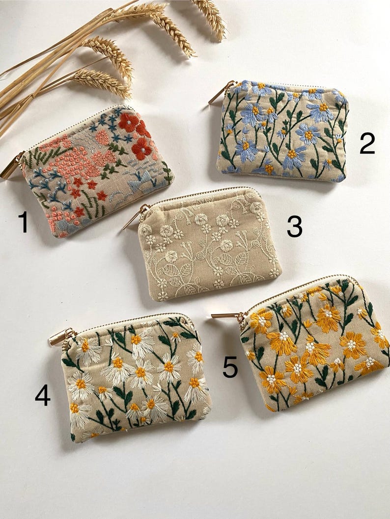 K&ouml;nnte beinhalten: F&uuml;nf kleine, beige bestickte M&uuml;nzb&ouml;rsen mit goldenen Rei&szlig;verschl&uuml;ssen. Die Geldb&ouml;rsen weisen florale Designs in verschiedenen Farben auf, darunter Rosa, Rot, Blau, Wei&szlig; und Gelb. Die Geldb&ouml;rsen sind auf einer wei&szlig;en Oberfl&auml;che mit Weizen&auml;hren im Hintergrund angeordnet.