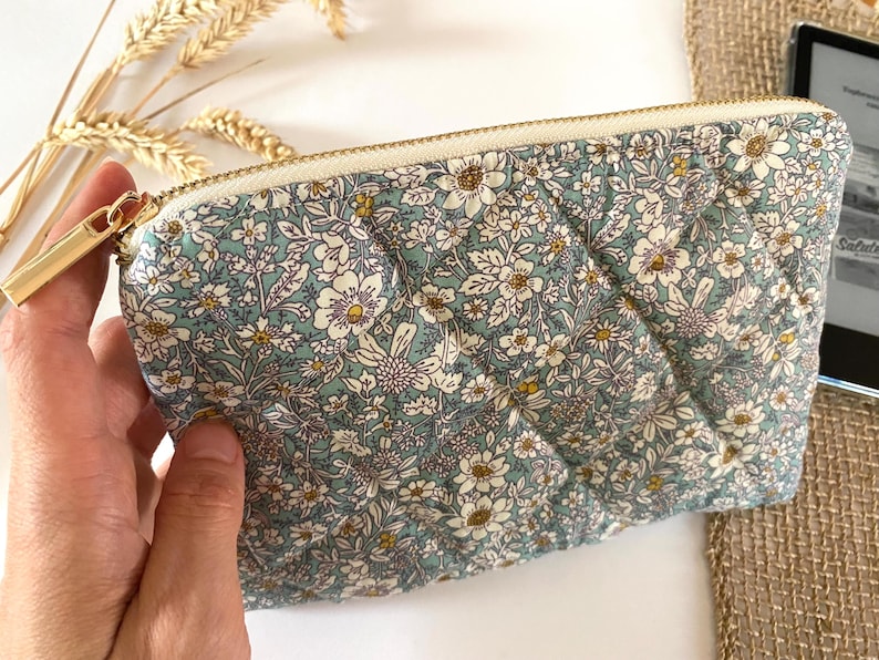 May include: A quilted pouch with a floral pattern in shades of blue, white, and yellow. The pouch has a gold zipper and a rectangular shape. The floral design is a repeating pattern of small flowers and leaves.
