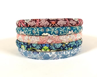 May include: A stack of five fabric-covered headbands, each featuring a distinct floral and leaf pattern. The colors include deep burgundy, teal, soft pink, navy blue, and light blue, all showcasing intricate botanical designs across the fabric.