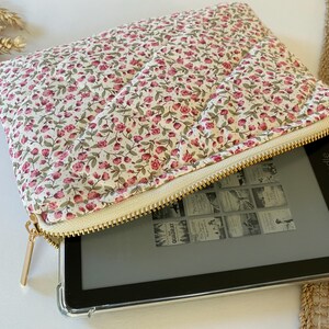 May include: A white and pink floral zippered pouch with a gold zipper. The pouch is open and a black Kindle e-reader is inside.