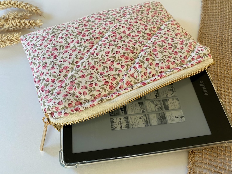May include: A white and pink floral zippered pouch with a gold zipper. The pouch is open and a black Kindle e-reader is inside.