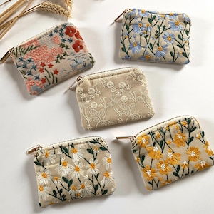 K&ouml;nnte beinhalten: F&uuml;nf kleine, rechteckige, bestickte M&uuml;nzb&ouml;rsen mit goldenen Rei&szlig;verschl&uuml;ssen. Die Geldb&ouml;rsen zeigen florale Designs in verschiedenen Farben, darunter Rosa, Blau, Wei&szlig; und Gelb, auf beigem Hintergrund. Die Stickereidetails umfassen G&auml;nsebl&uuml;mchen und andere botanische Motive.