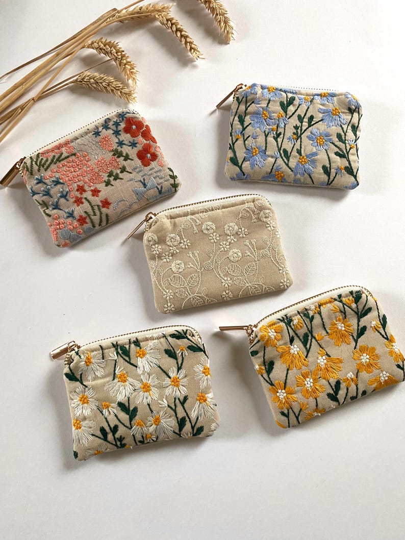 K&ouml;nnte beinhalten: F&uuml;nf kleine, rechteckige, bestickte M&uuml;nzb&ouml;rsen mit goldenen Rei&szlig;verschl&uuml;ssen. Die Geldb&ouml;rsen zeigen florale Designs in verschiedenen Farben, darunter Rosa, Blau, Wei&szlig; und Gelb, auf beigem Hintergrund. Die Stickereidetails umfassen G&auml;nsebl&uuml;mchen und andere botanische Motive.