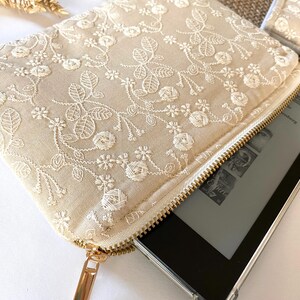 May include: A beige clutch with a floral lace overlay and a gold zipper. The clutch is open, revealing a black e-reader. The lace design features detailed floral and leaf patterns. A second clutch is visible in the background.
