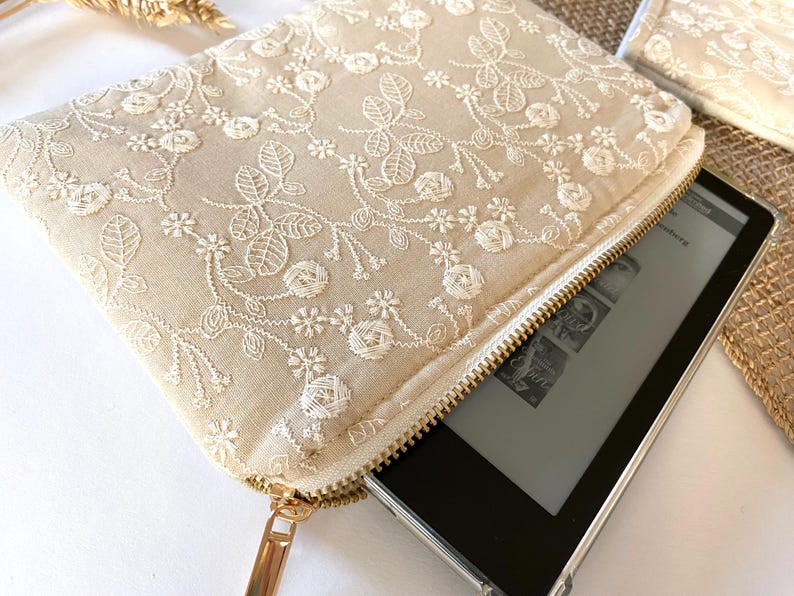 May include: A beige clutch with a floral lace overlay and a gold zipper. The clutch is open, revealing a black e-reader. The lace design features detailed floral and leaf patterns. A second clutch is visible in the background.