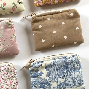 May include: A collection of small, zippered pouches in various floral and patterned fabrics. One pouch is tan with white hearts, and another features a blue and white toile design. The pouches have gold-toned zippers.