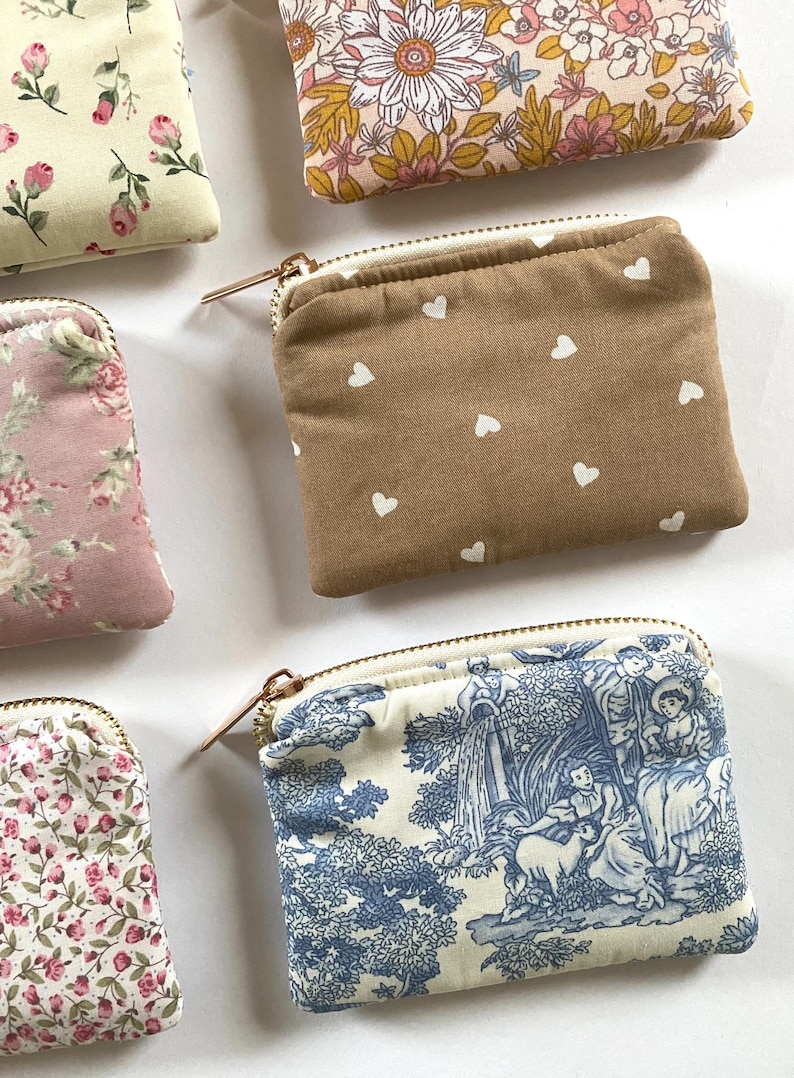 May include: A collection of small, zippered pouches in various floral and patterned fabrics. One pouch is tan with white hearts, and another features a blue and white toile design. The pouches have gold-toned zippers.