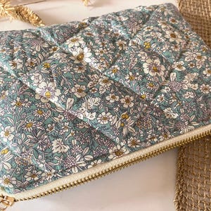 May include: A quilted floral clutch in shades of teal, white, and yellow. The rectangular pouch features a repeating flower pattern and a gold-toned zipper closure. The fabric has a diamond-shaped quilted design.